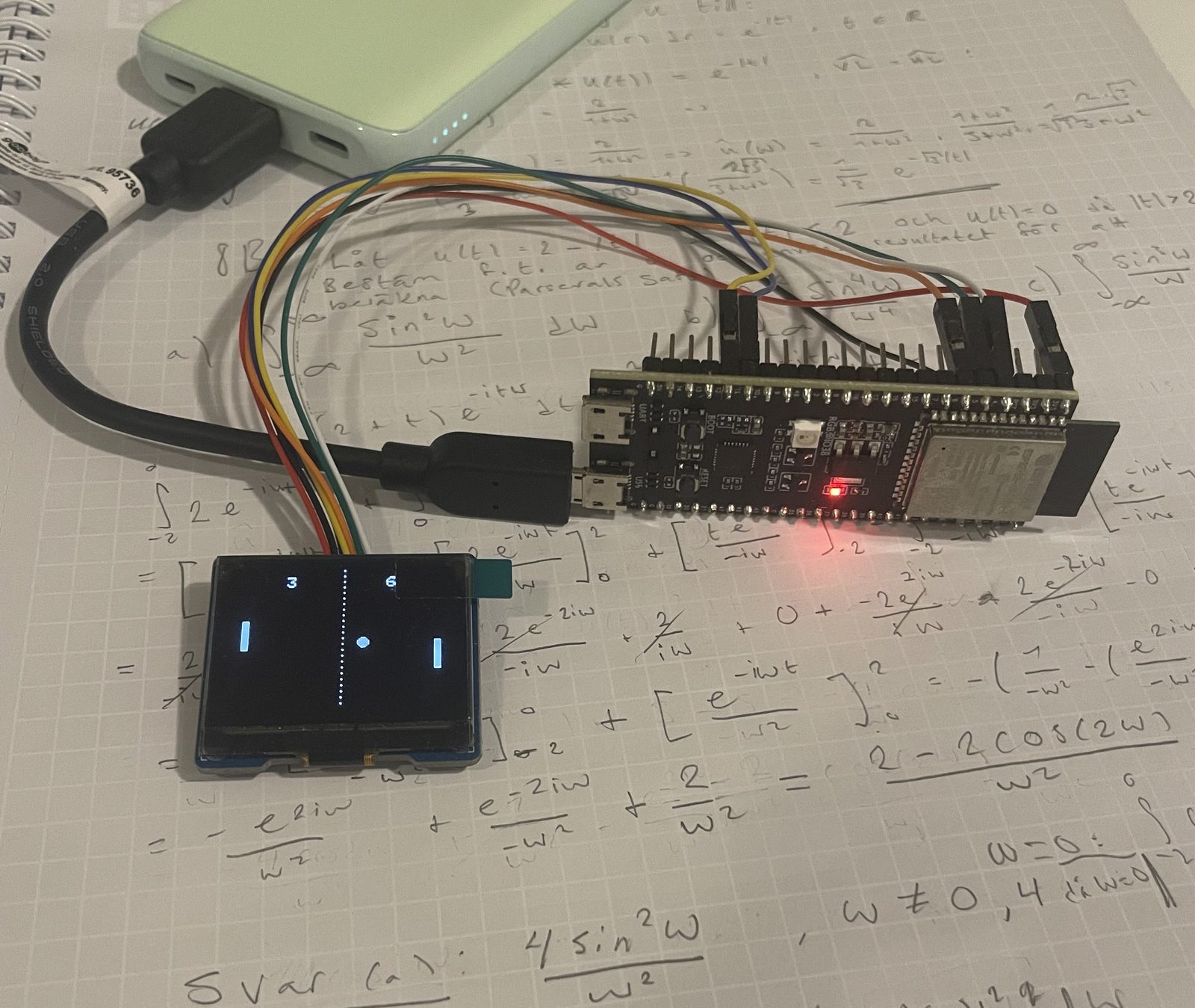 An ESP32-S3 development board wired to a small OLED display, resting on a page of handwritten Fourier analysis