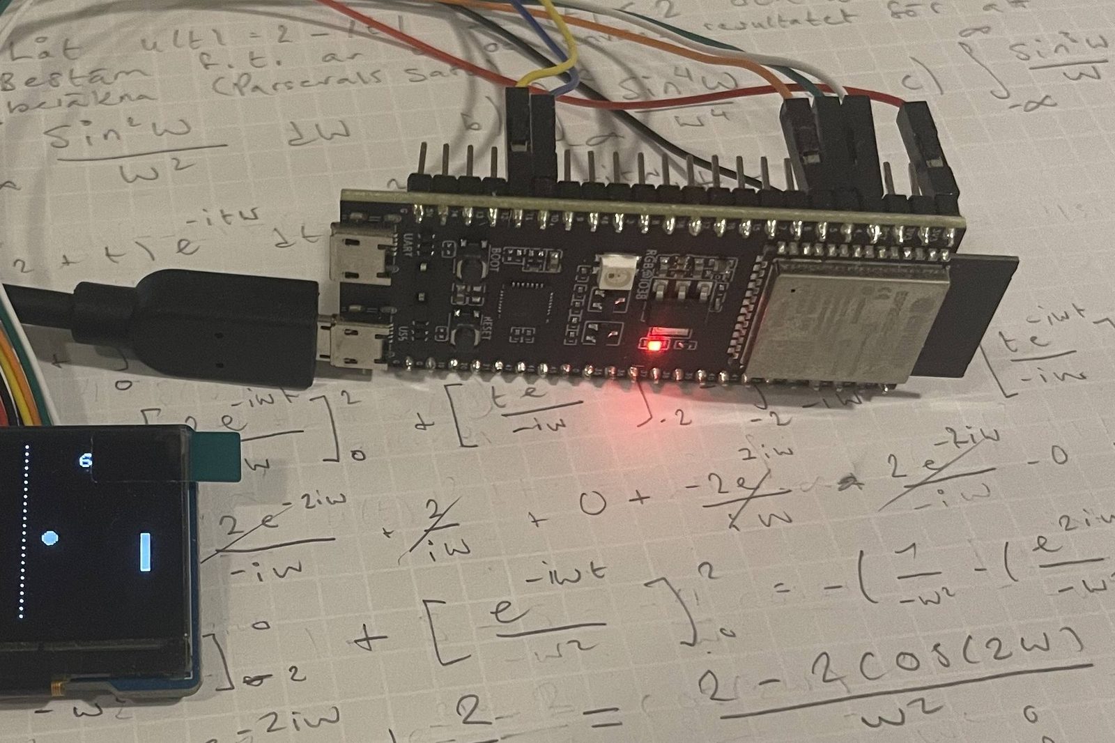 An ESP32-S3 board and a small OLED display running on a page of handwritten Fourier analysis